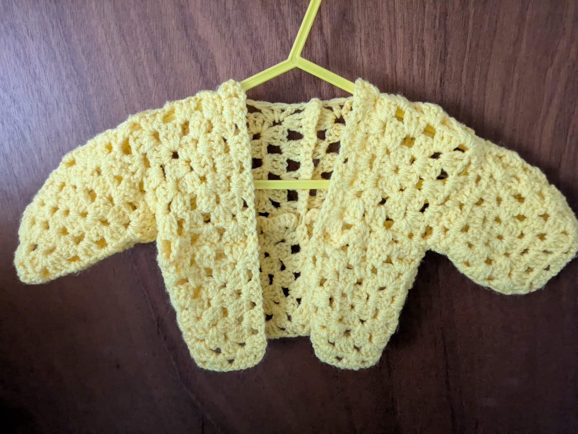 Yellow crocheted baby cardigan on a hanger against a wooden background.