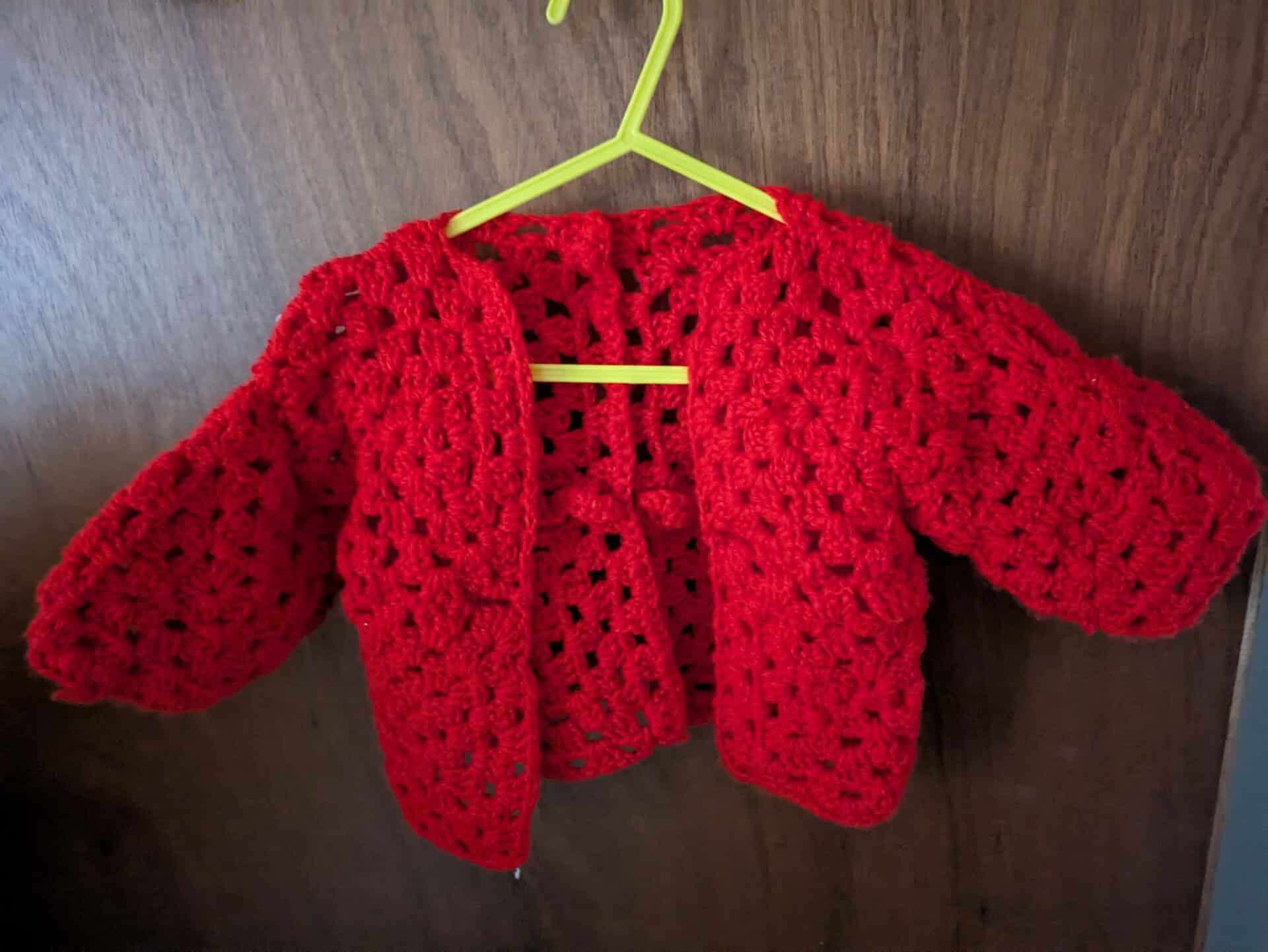 Red crocheted baby cardigan on a yellow hanger.