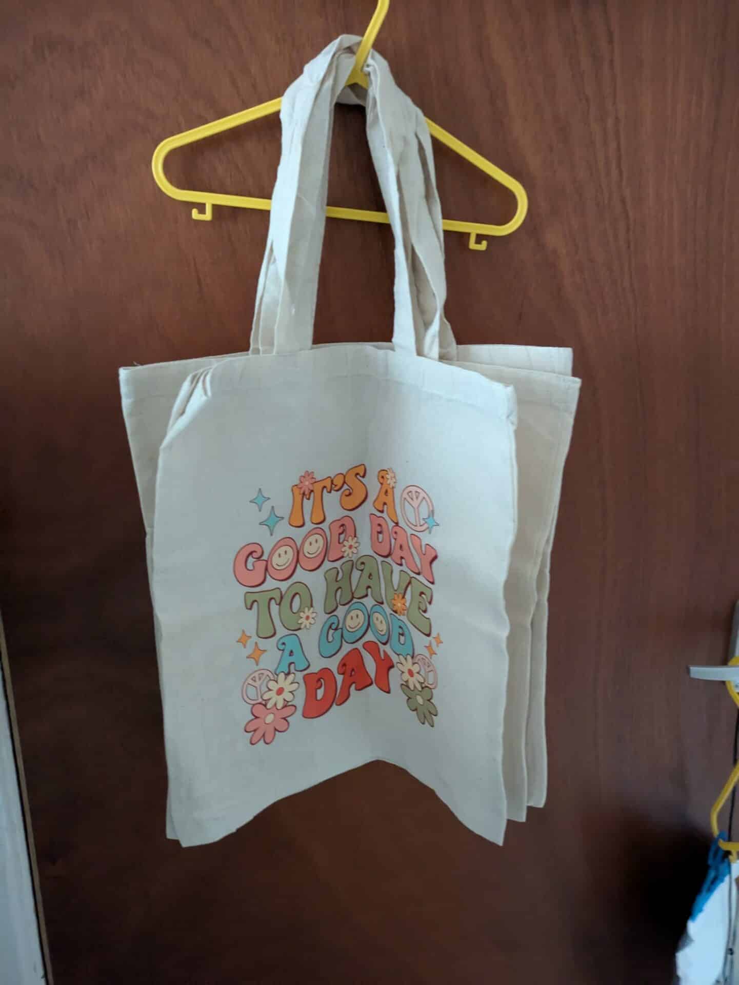 Canvas tote bag with colorful text, "It's a good day to have a good day," hanging on a yellow hanger.
