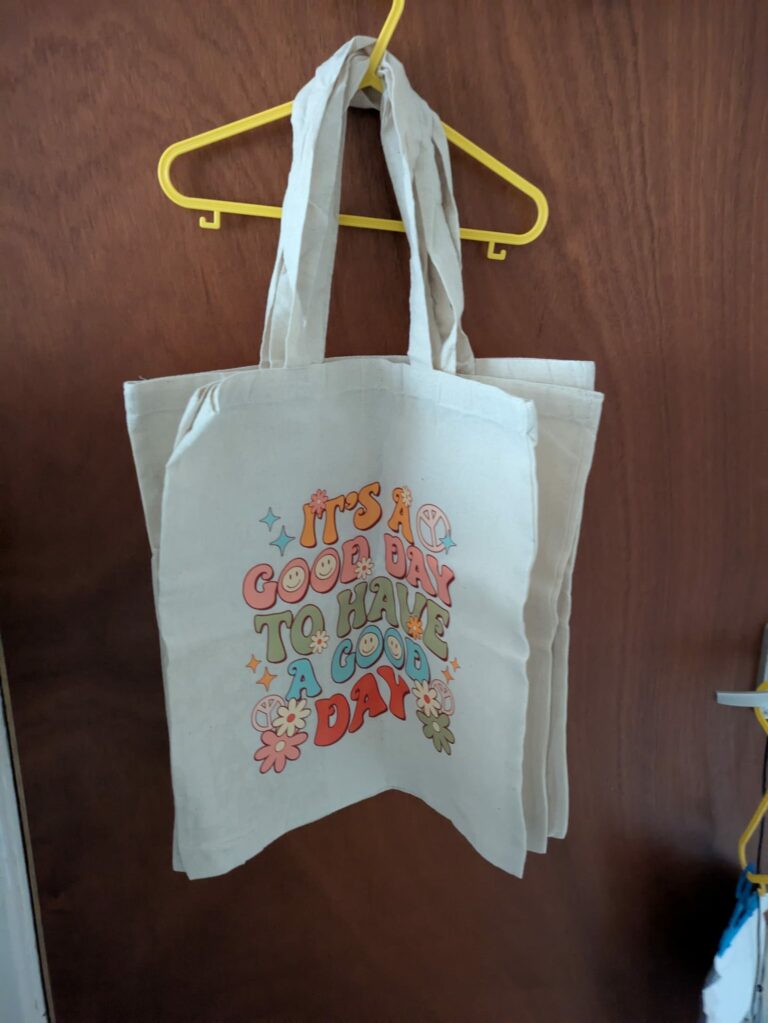 Canvas tote bag with colorful text, "It's a good day to have a good day," hanging on a yellow hanger.