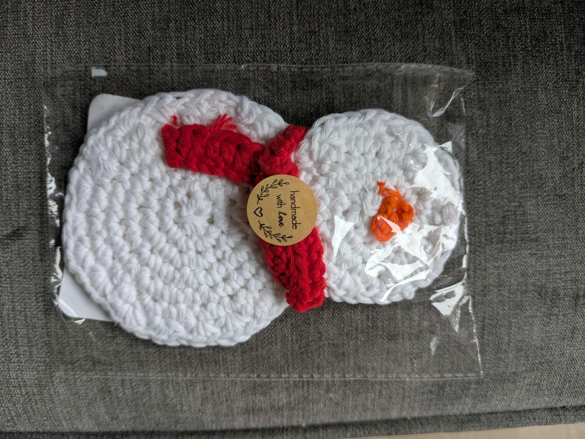 Crocheted snowman with red scarf, packaged in clear plastic, on a textured gray surface.