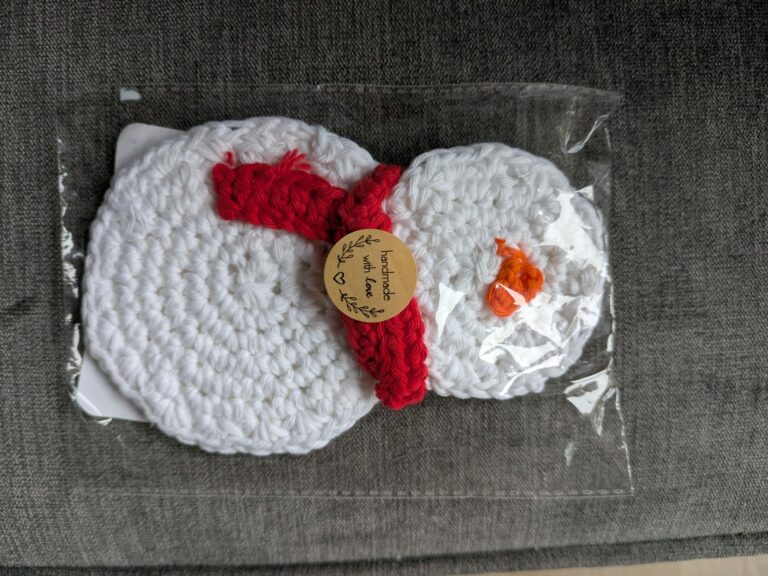 Crocheted snowman with red scarf, packaged in clear plastic, on a textured gray surface.