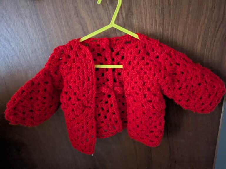 Red crocheted baby cardigan on a yellow hanger.