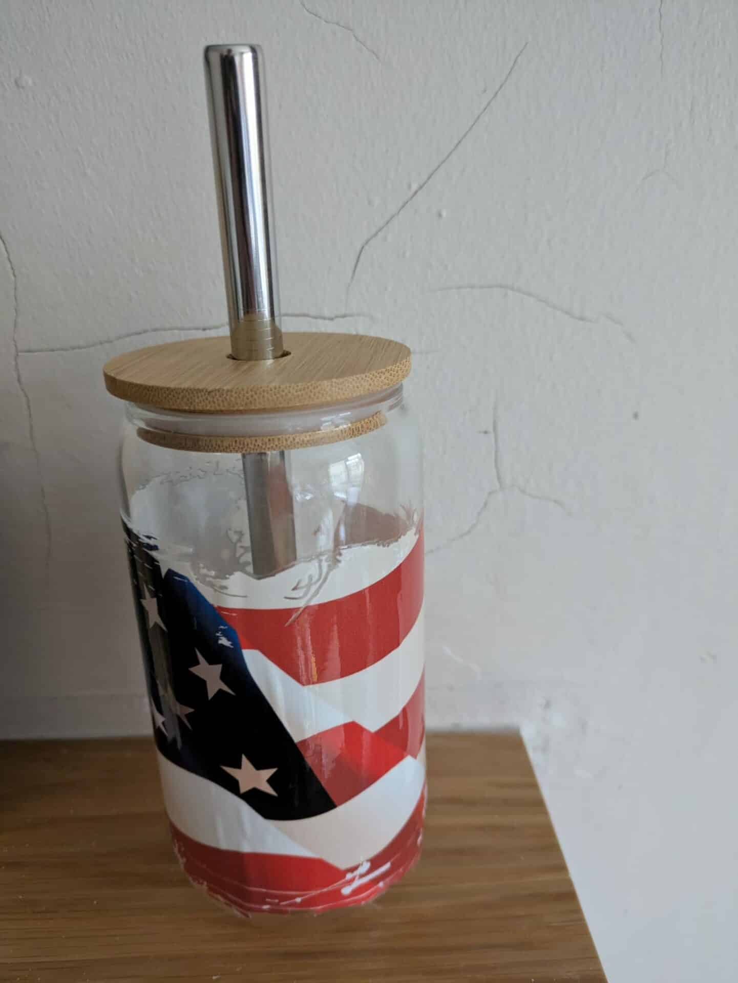 Glass tumbler with American flag design, bamboo lid, and metal straw on a wooden surface.