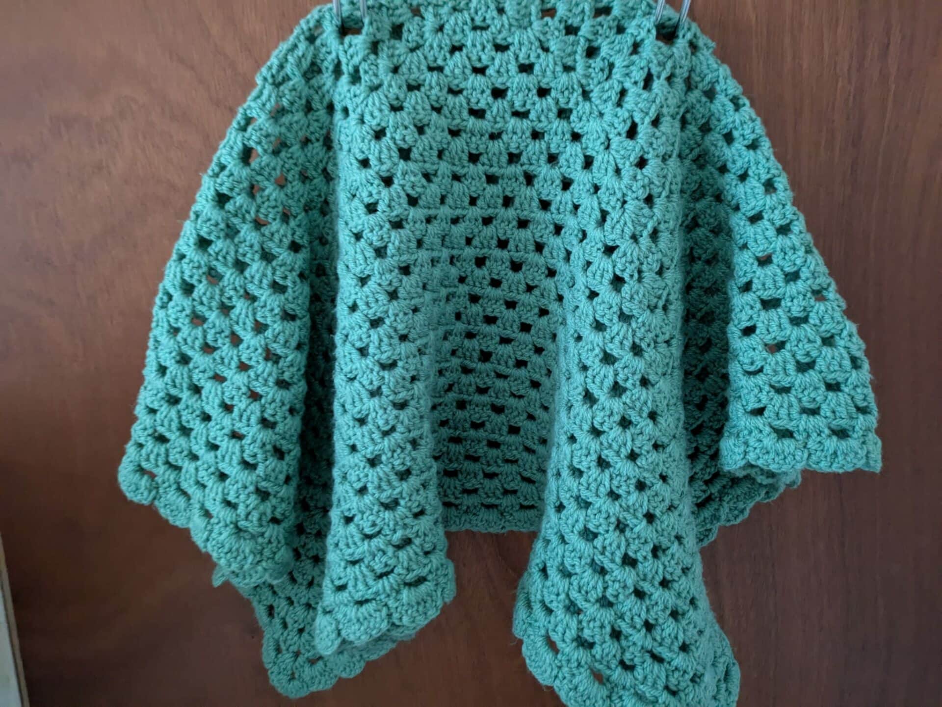 Green crocheted shawl hanging against a wooden background.