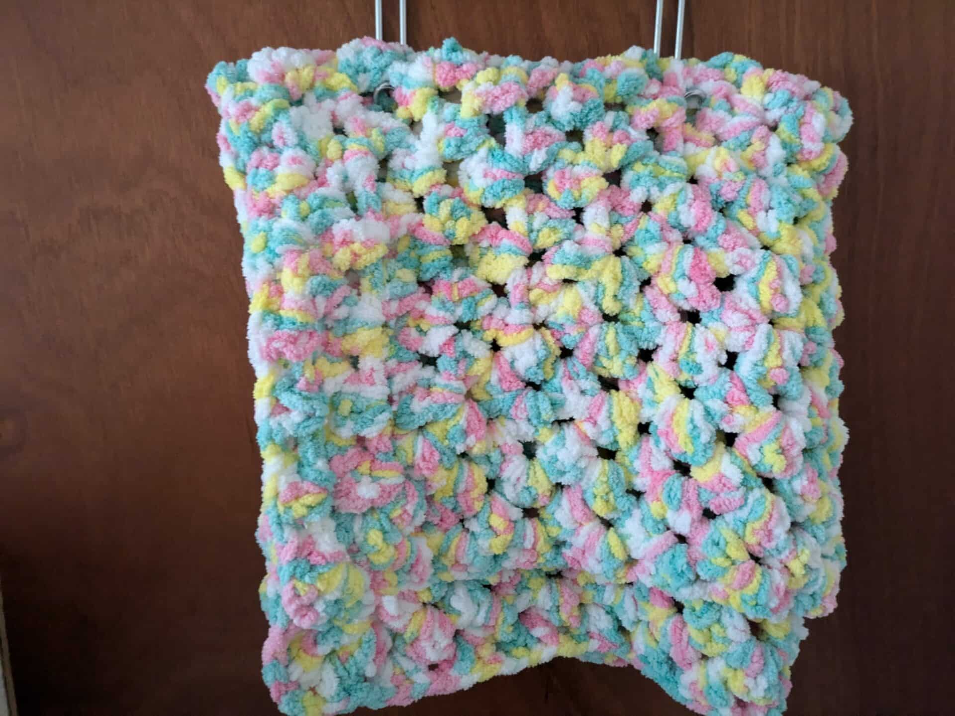Colorful crochet square with pink, yellow, blue, and white yarn hanging on a wooden background.