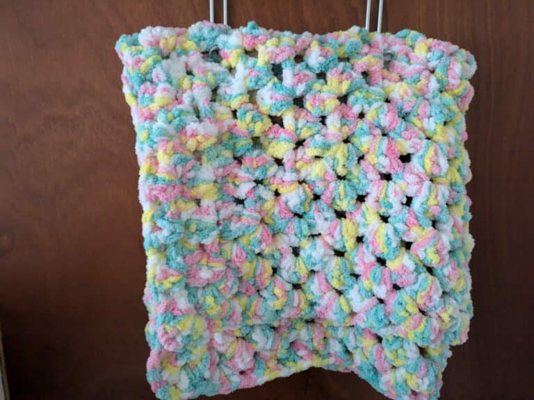 Colorful crochet square with pink, yellow, blue, and white yarn hanging on a wooden background.