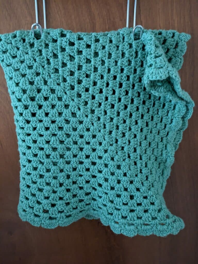 Green crocheted granny square blanket hanging on hooks against a wooden background.