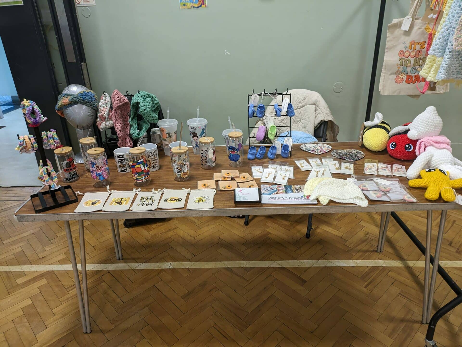 Craft fair display featuring crocheted items, decorated cups, keychains, and small plush toys on a wooden table.